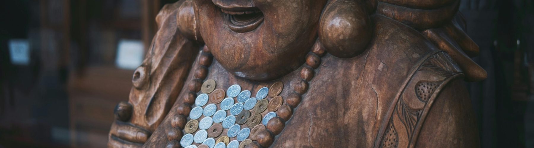 Wooden sculpture of a laughing Buddha adorned with coins and beads, showcasing spiritual abundance.